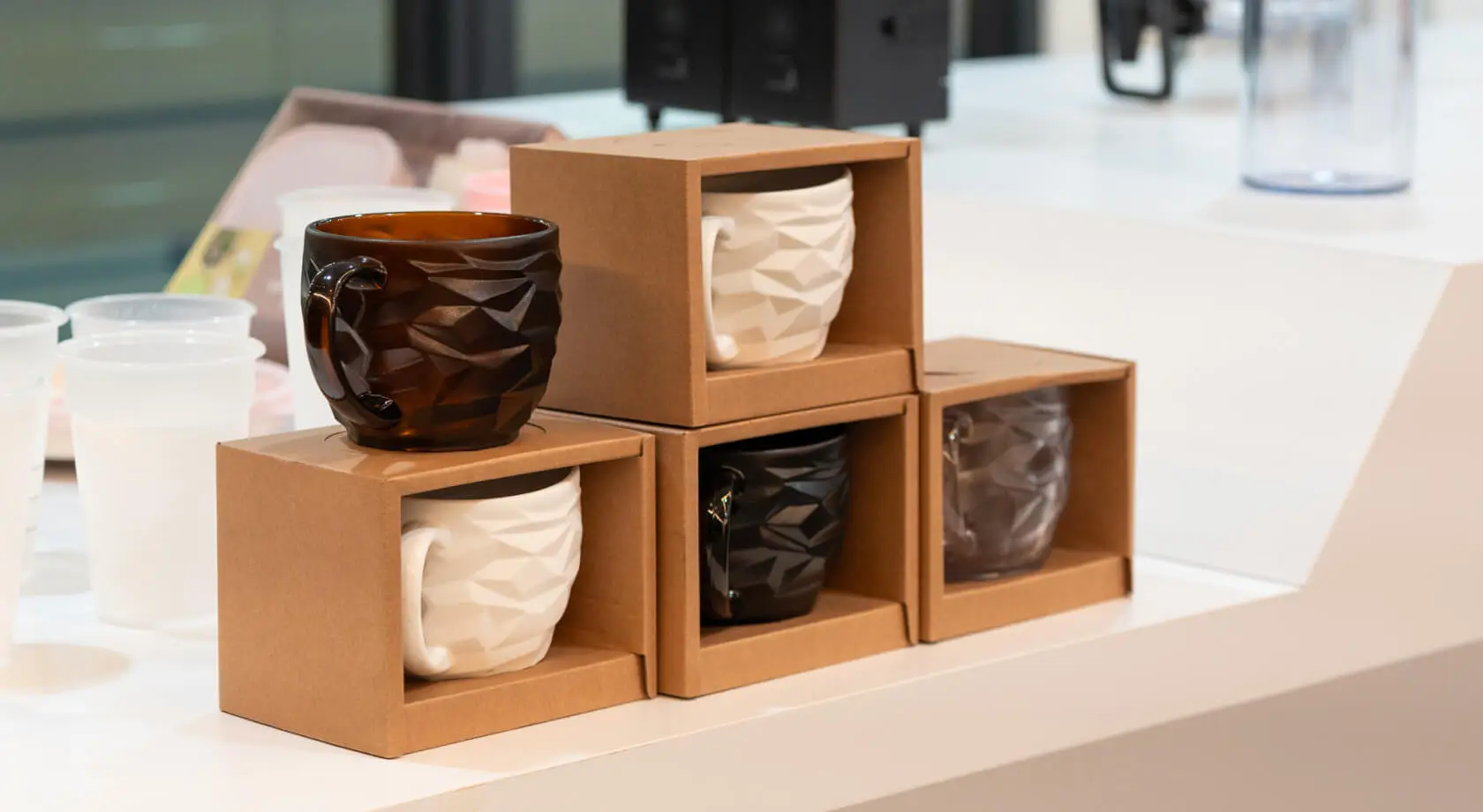 Injection-molded faceted drinkware mugs in white, black, smoke, and amber finishes displayed in cardboard packaging at an AMP showroom