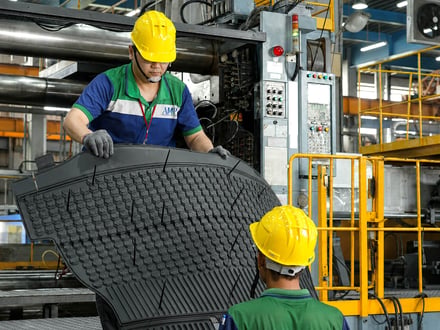 AMP operators removing a large injection-molded automotive floor mat directly from the press, showing the textured surface detail produced in a single shot